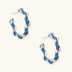 White gold-plated hoops intertwined with blue denim fabric in a twisted rope design on cream background.