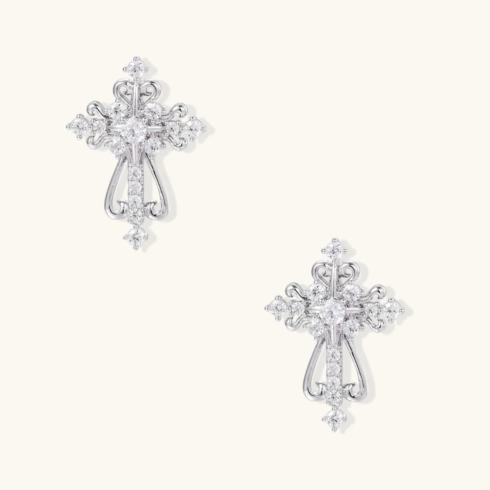 White gold-plated baroque cross studs with zirconia stones and ornate scrollwork on cream background.