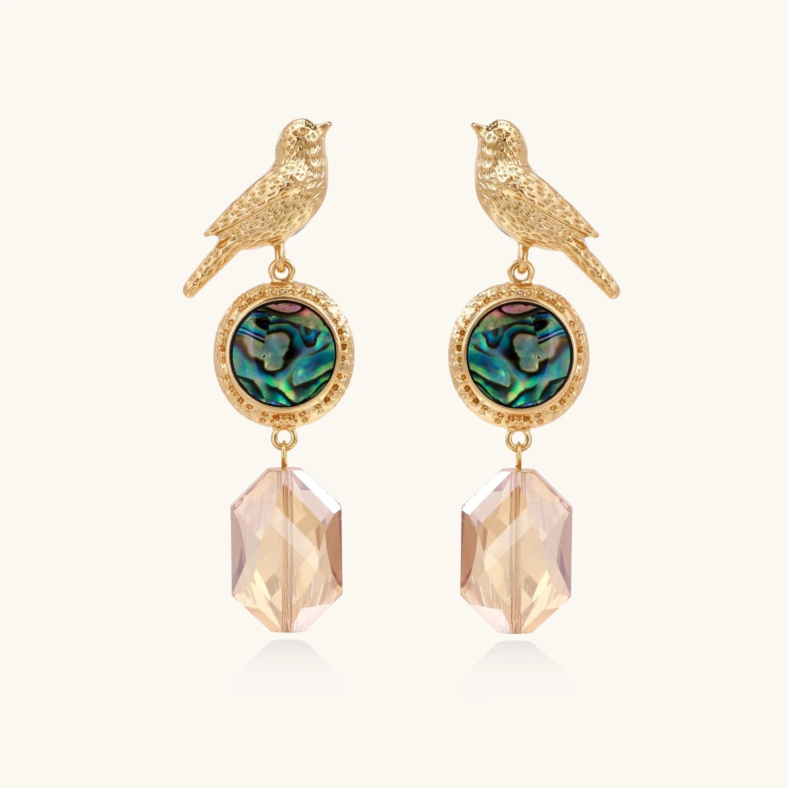 Gold-plated bird stud earrings with iridescent abalone shell medallions and champagne crystal drops on cream background
