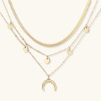 Four-layer gold necklace with herringbone chain, disc charms, and crescent moon pendant on cream background