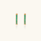 Pair of small gold huggie hoop earrings with bright green cubic zirconia stones on cream background