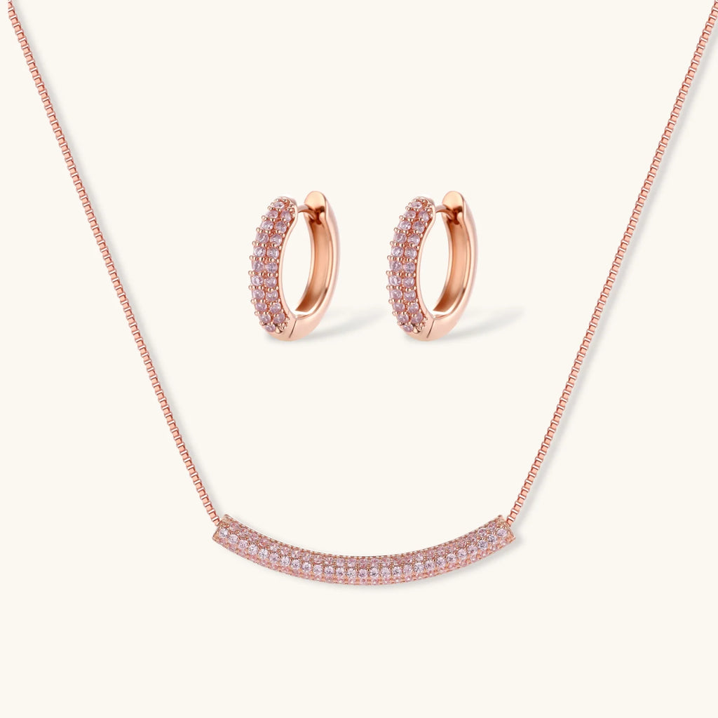Rose gold jewelry set with pavé cubic zirconia stones including curved necklace, hoop earrings, and adjustable ring on cream background.