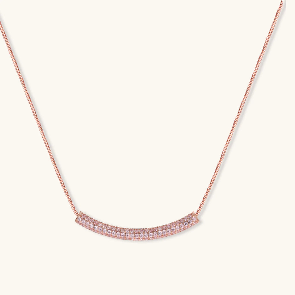 Rose gold jewelry set with pavé cubic zirconia stones including curved necklace, hoop earrings, and adjustable ring on cream background.