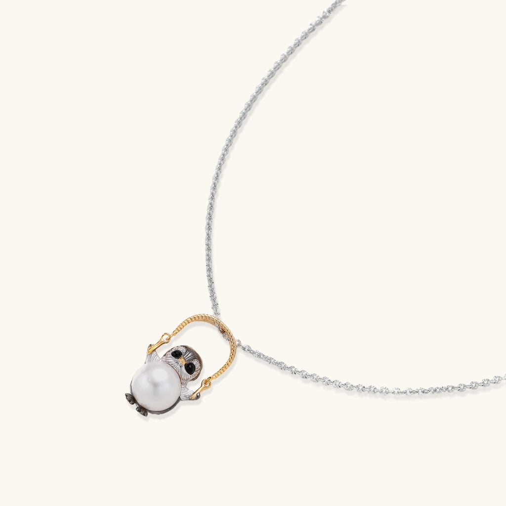 Whimsical owl pendant holding a white pearl on a gold swing, rhodium-plated silver chain on cream background.
